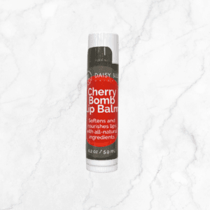 A tube of lip balm with a red label stands upright on a white marble surface next to Flawless Glow Blush, highlighting its all-natural ingredients and net weight of 0.2 oz (5.9 ml).