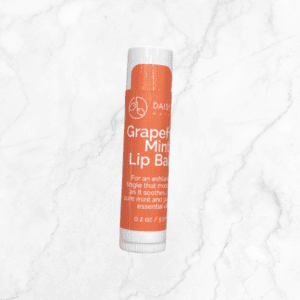 A tube of Daisy Naturals Grapefruit Mint Lip Balm with an orange label stands upright on white marble. Pair it with Flawless Glow Blush for radiant skin, while the lip balm soothes with pure mint and grapefruit oils.