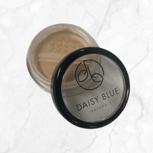 A small round container of Magnolia Foundation loose powder, with a black-rimmed clear lid partially open, rests elegantly on a white marble surface.