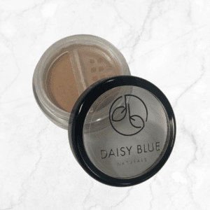 An open container of Mahogany Foundation loose powder sits on a white marble surface. The beige powder shows through a clear lid with a black rim, which displays the Mahogany Foundation logo and product name.