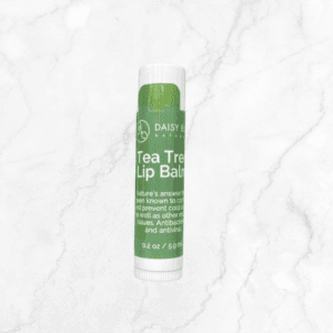 A tube of Daisy Nature Tea Tree Lip Balm stands upright on white marble, next to Flawless Glow Blush. The green and white lip balm label shows product details and a net weight of 0.2 oz (5.9 ml).