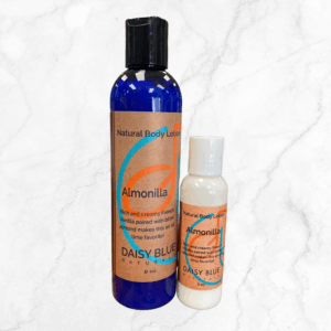Two bottles of Almonilla Lotion are shown on a white marble background. The larger blue bottle with a black cap and the smaller white bottle have brown labels, both showcasing the signature Almonilla scent.