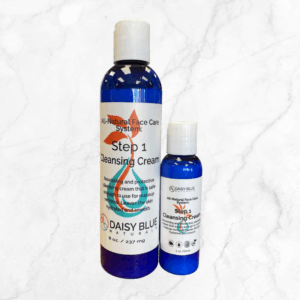 Two blue bottles of Cleansing Cream, one large and one small, sit on a light marble background. Both have white caps and labels describing the product.