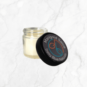 A small glass jar of Marigold Salve with a cream hue and black lid, featuring the "Daisy Blue Naturals" logo, rests on a light marble background.