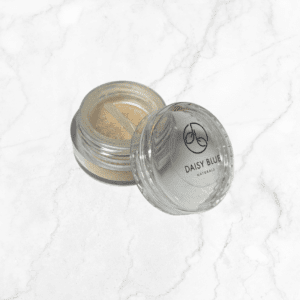An open jar of Powderflauge Concealer - LIGHT loose powder sits on a white marble surface. The small clear container with a sifter lid features "DAISY BLUE NATURALS" and its logo printed on the top.