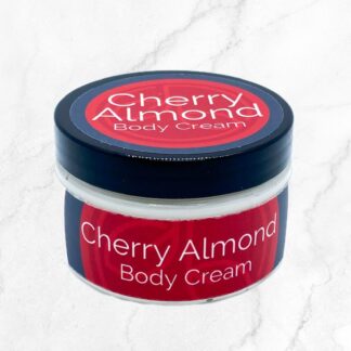 A small round jar with a black lid labeled "Cherry Almond Body Cream" rests on white marble, offering rich hydration and a sweet cherry almond scent.