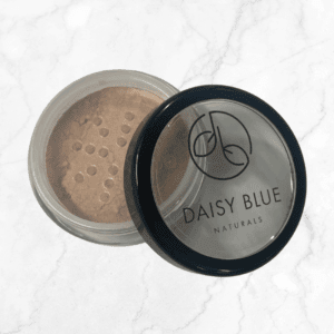 A partially opened Poplar Foundation loose powder makeup container with a black lid labeled "Daisy Blue Naturals" sits on white marble, showing tan powder visible through the sifter inside.