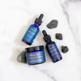 The Elite Skin Care Trio—two "Liquid Gold" dropper bottles and one "Whipped C Daily Defense" jar—are beautifully arranged on white marble with black stones, highlighting this luxurious set.