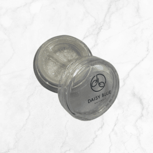 A compact, round clear container of Stardust Eyeshadow features a split compartment with shimmery powder. The lid displays "DAISY BLUE NATURALS" and logo, and the container sits elegantly on a white marble surface.