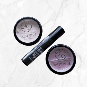The Mineral Makeup Starter Trio, featuring two round containers in light and rosy shades plus a mascara tube, is arranged on white marble. Its translucent lids display the colors, making this an ideal starter kit for mineral makeup.