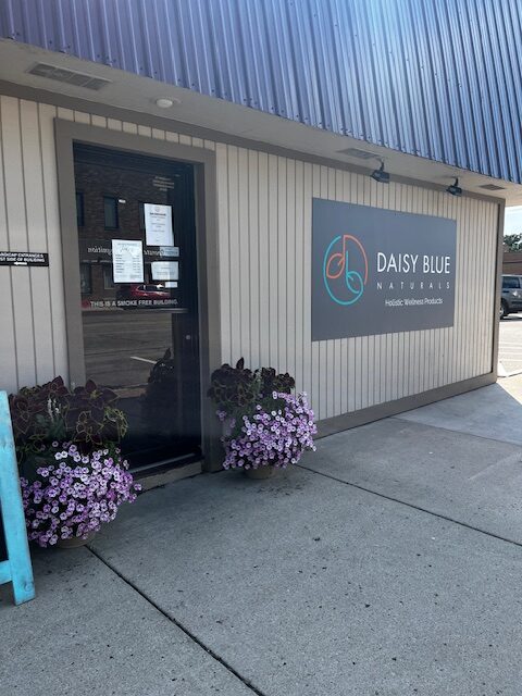 Exterior of Daisy Blue Naturals in Owatonna, MN, with a large sign on the building. The entrance features signs on the glass door and two planters with purple flowers flanking the doorway.