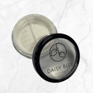 A clear round container with a black lid labeled "DAISY BLUE NATURALS" sits on a white marble surface, partially open to show the Matte Cream Primer’s fine white texture inside.