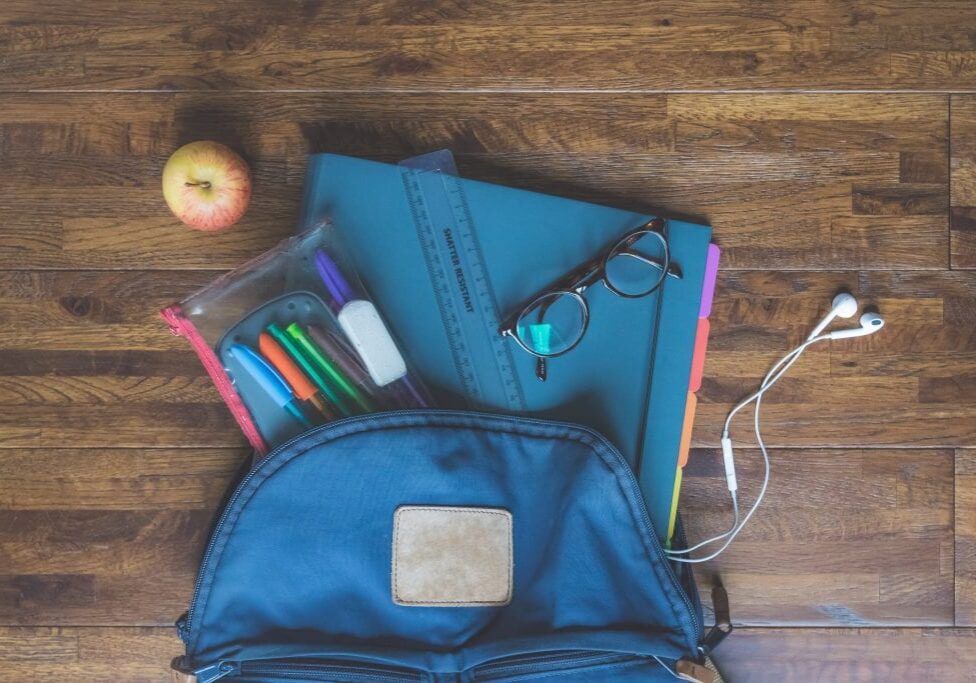 A perfect Back to School essential: a blue backpack on a wooden floor with an apple, folders, a ruler, eyeglasses, earphones, and a clear pouch with markers and an eraser partially spilling out.