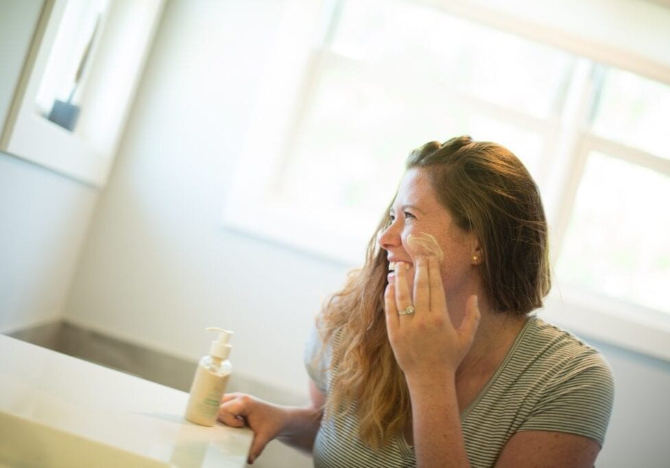 A woman with long hair applies cream to her face while smiling in a bright bathroom, confidently caring for her skin type, with a bottle of lotion on the counter in front of her.