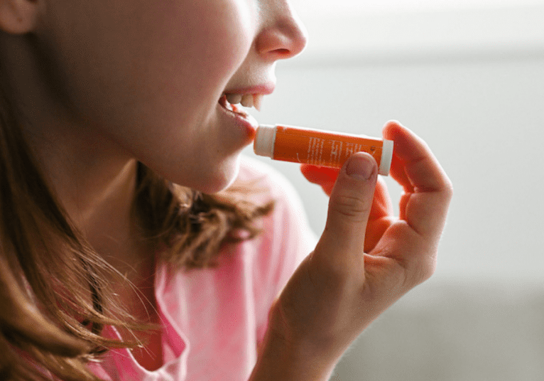 A child wearing a pink shirt applies Daisy Blue natural lip balm to their lips, holding the tube with one hand. The background is softly blurred.