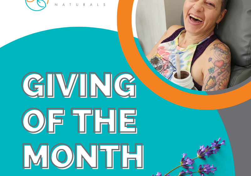 A smiling woman with tattoos sits in a chair holding a cup. The text reads “Giving of the Month,” celebrating natural skincare & wellness, with a purple flower and the Daisy Blue Naturals logo above.