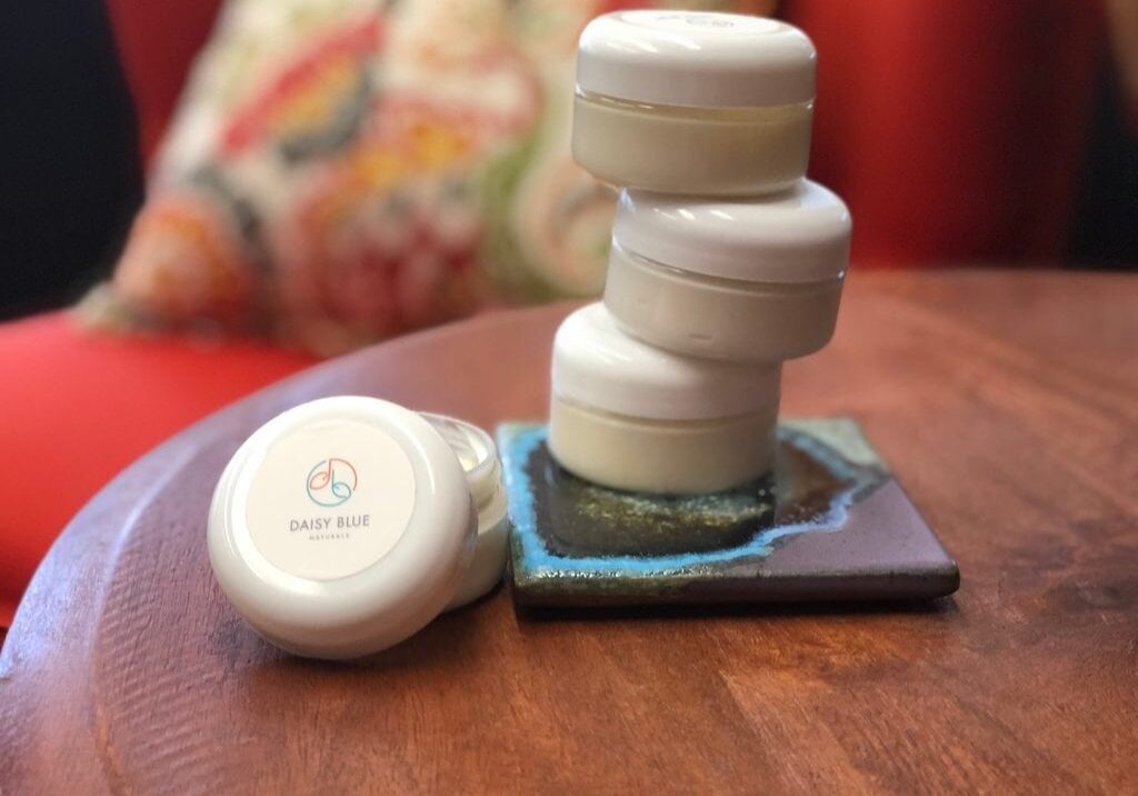 Four small white jars of Daisy Blue body butters, three stacked and one leaning, rest on a wooden table beside a decorative square tile. The colorful, blurred background hints at the nourishment these jars bring.
