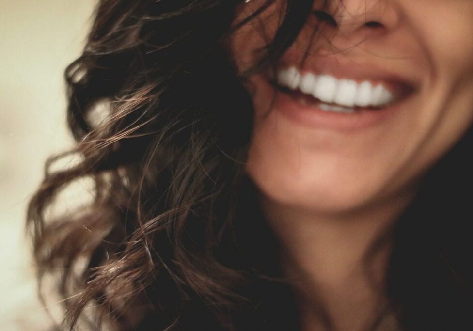 Close-up of a person with wavy dark hair smiling, showing white teeth. Though their eyes are hidden, the joyful mood captures how they age beautifully—radiating carefree charm like a Daisy Blue moment.