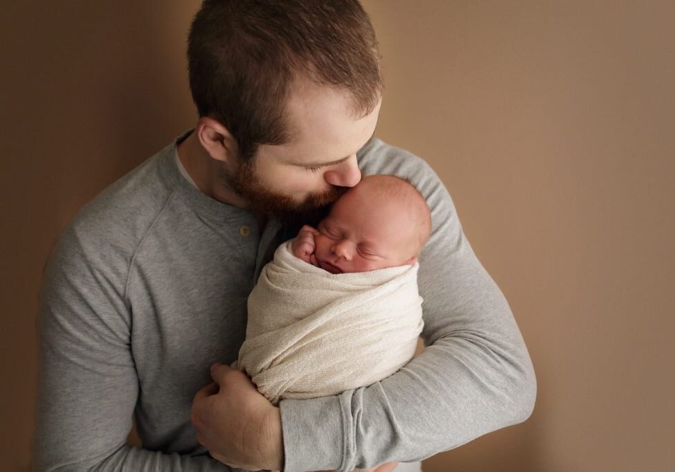 A man in a gray shirt gently holds and kisses a sleeping newborn wrapped in a cream blanket against a soft brown background, capturing the perfect happy fathers day moment.