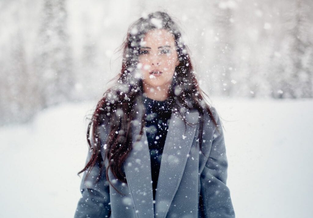A woman with long dark hair wearing a gray coat stands in a snowy landscape, embodying the Winter Blues as snow falls around her, partially obscuring her face and body. The background is blurred with snow-covered trees.