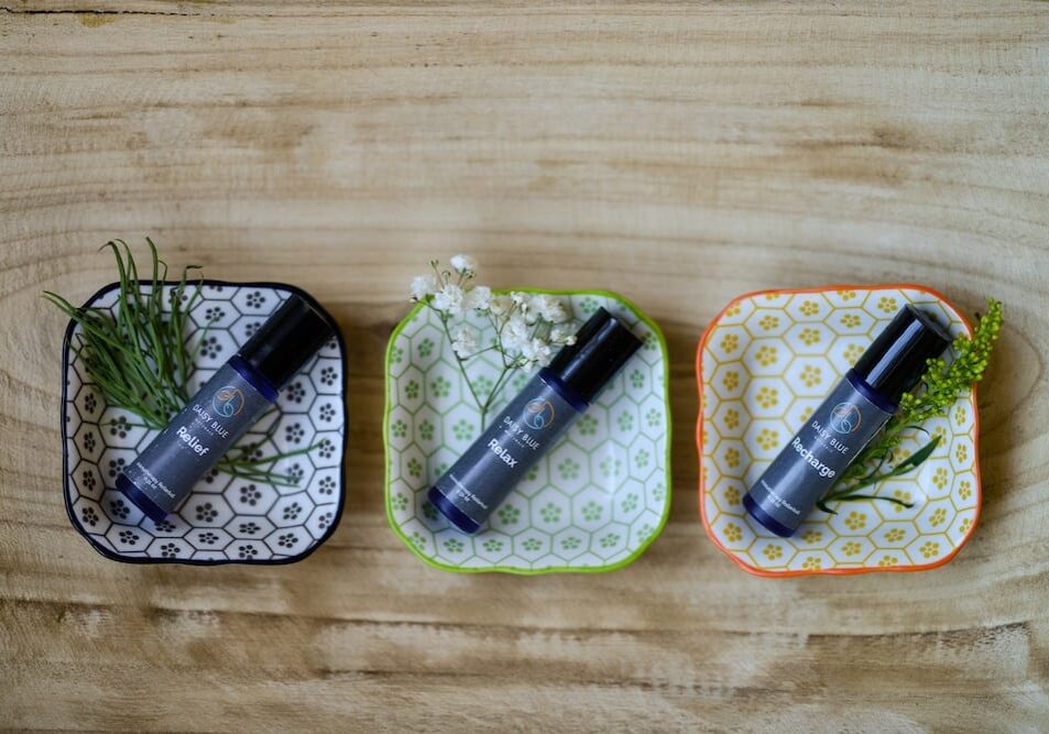 Three small decorative plates, each holding a blue Essential Oil Rollerball and a sprig of greenery or flowers, are arranged in a row on a light wooden surface.
