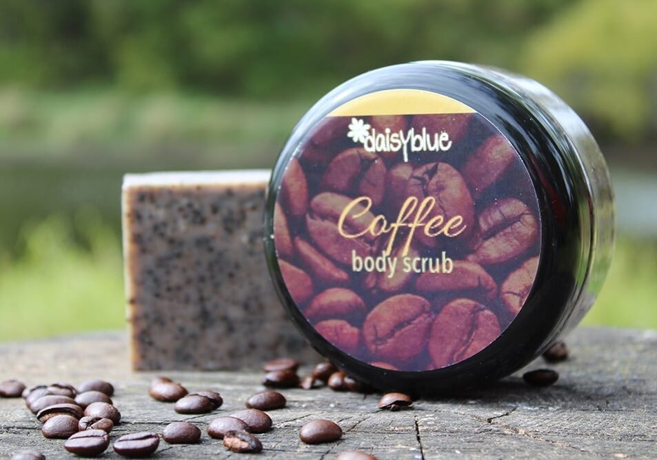 A jar labeled "daisyblue Coffee body scrub" sits on a wooden surface next to a bar of scrub soap and scattered coffee beans, highlighting the health benefits of coffee, with blurred greenery in the background.