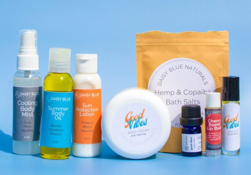 A lineup of Daisy Blue Naturals skincare products from the Good Vibes Box, including body mist, lotion, bath salts, and more, displayed on a blue background.