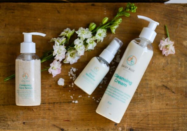 Three skincare products labeled “Cleansing Face Scrub,” “Facial Moisturizer,” and “Cleansing Cream” are arranged on a wooden surface with white flowers and scattered grains of salt, ideal to prep skin for makeup and perfect your skin preparation routine.