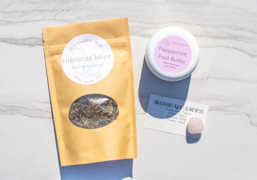 A Limited-Edition May Trio: a packet of hibiscus mint tea, peppermint foot butter, a rose quartz crystal, and an info card are artfully arranged on a light marble surface.
