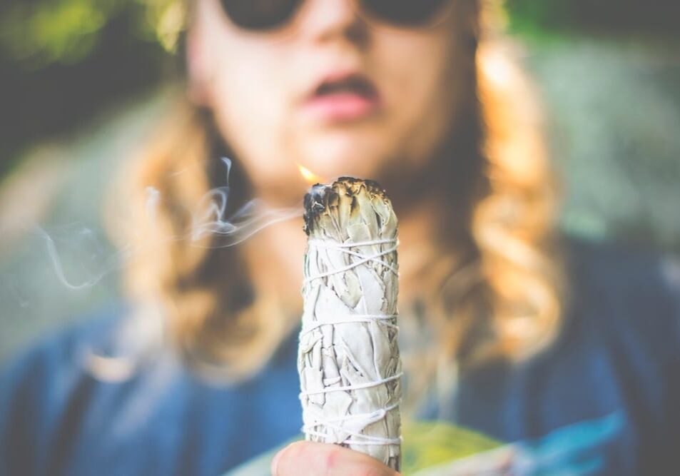 A person with long blond hair and sunglasses holds a smoking bundle of sage, wrapped in white string, in front of them, practicing smudging for cleansing. The background is blurred.