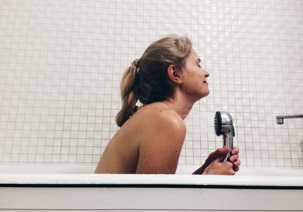 A woman with blonde hair in a ponytail sits in a white bathtub filled with bubbles, holding a shower head and smiling, enjoying some relaxing hair care, with a tiled white wall in the background.