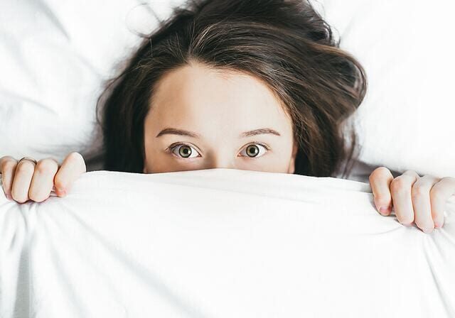 A person with long brown hair lies in bed, holding a white blanket up to their nose, only their wide eyes and forehead visible—perhaps contemplating skincare or if it's okay to sleep with makeup as they gaze directly at the camera.