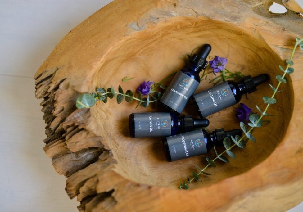 Four small dropper bottles labeled "Peppermint Oil" and "Lavender Oil" rest inside a large, rustic wooden bowl with green sprigs and purple flowers, creating an inviting setting for Holiday Wellness with pure essential oils.