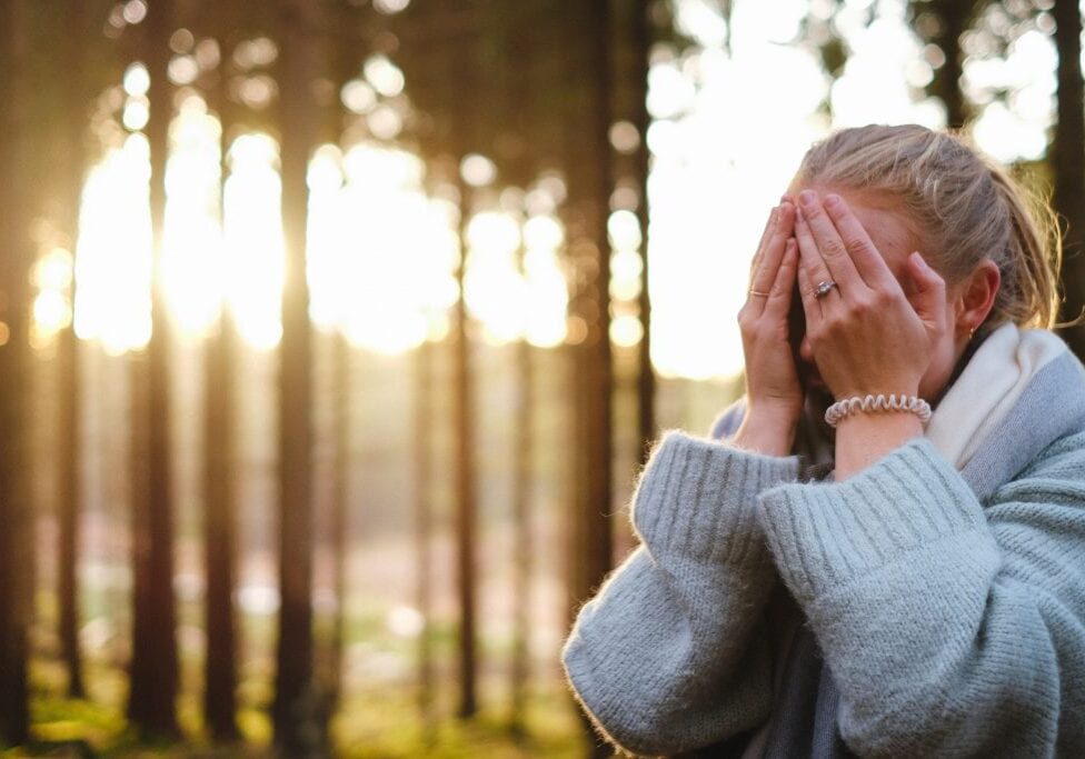 A person wearing a blue sweater and scarf stands in a sunlit forest, covering their face with both hands—perhaps to protect their skin or showcase the importance of face care as sunlight filters through the tall trees in the background.