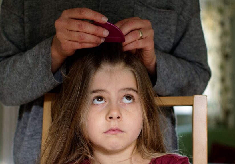A young girl with long brown hair sits on a chair, looking up with a bored expression as an adult uses a pink comb for simple lice removal. The adult’s hands are visible, but their face is out of frame.