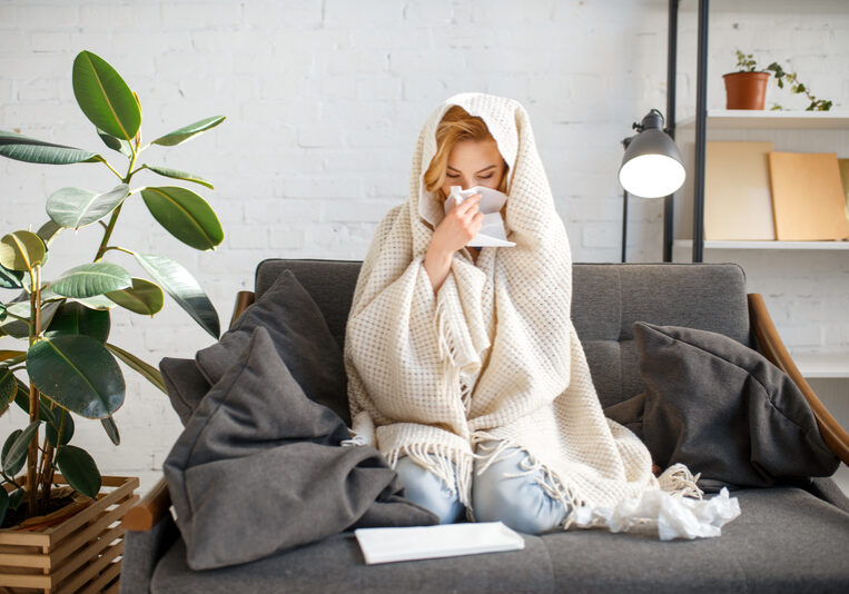 A person sits on a couch wrapped in a blanket, holding a tissue to their nose. Used tissues hint at cold and flu season. Nearby, a large plant and lamp create a cozy scene as they try to beat the cold with rest and comfort.