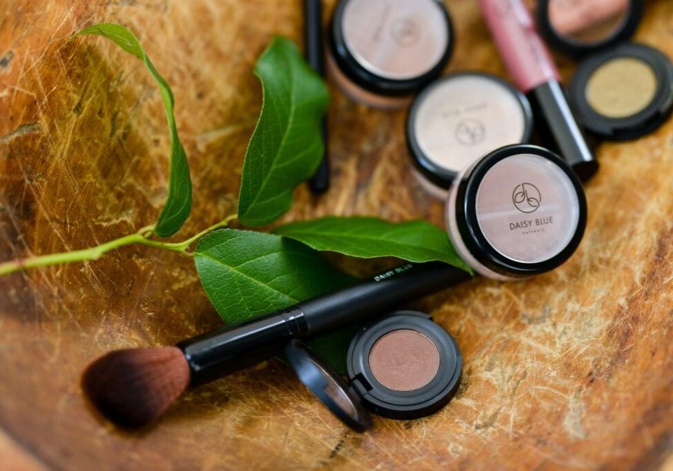 A close-up of Daisy Blue cosmetics, highlighted by jars and a makeup brush, sits alongside a green leafy branch on textured wood—reflecting the trust seen in our Lifetime Testimonials.
