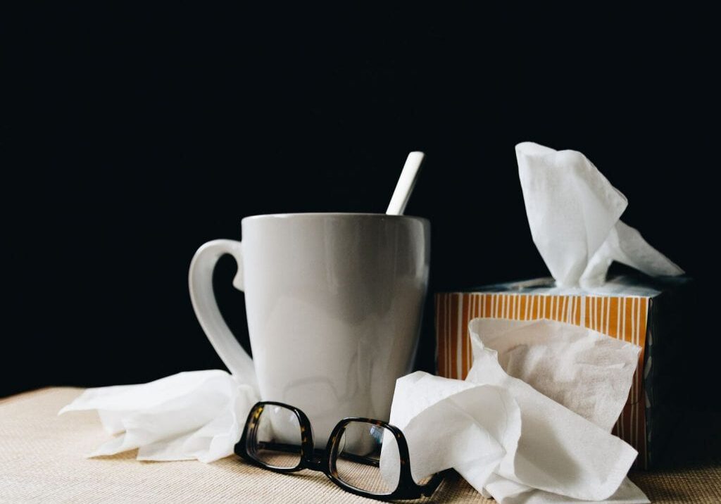 A white mug with a spoon inside sits next to a striped tissue box, used tissues, and a pair of black-rimmed glasses—essentials for natural cold remedies—on a light surface against a dark background.