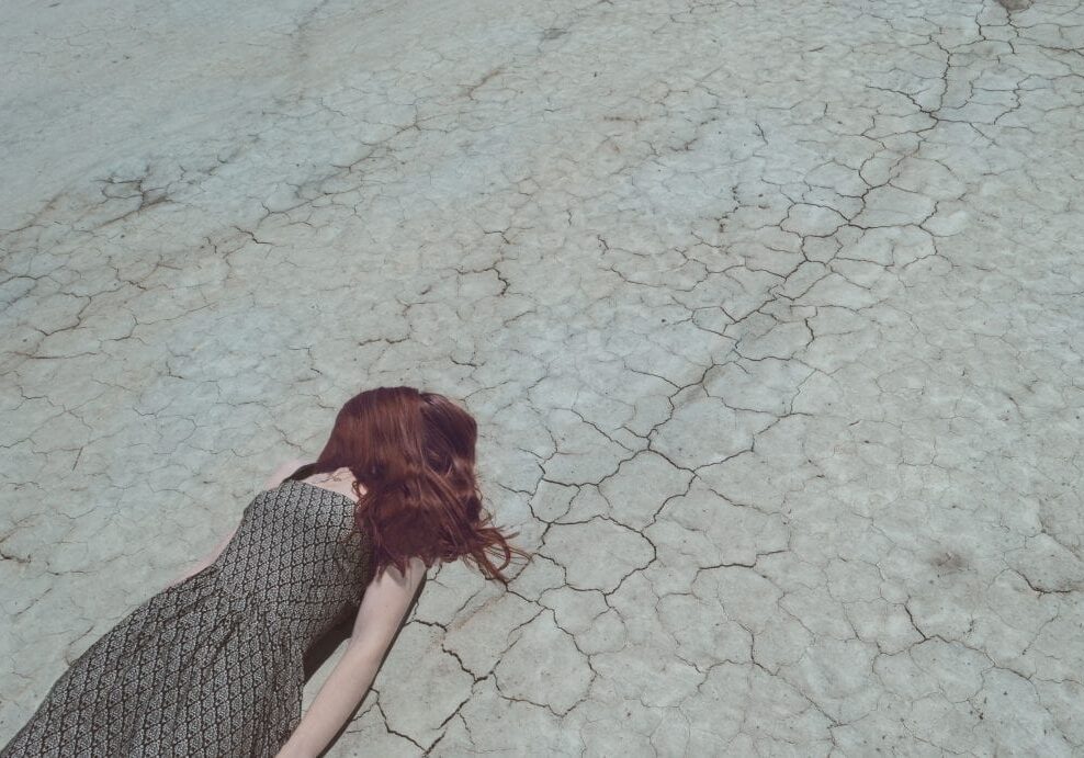 A woman with long red hair wearing a patterned dress lies face down on cracked, dry ground, suggesting a barren or drought-stricken landscape in need of natural eczema relief.