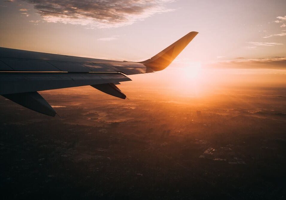 Airplane wing seen from a passenger window during flight, with a golden sunrise or sunset casting warm light over the landscape and clouds below—a perfect moment to reflect on travel tips or refresh your natural skincare routine while in the air.