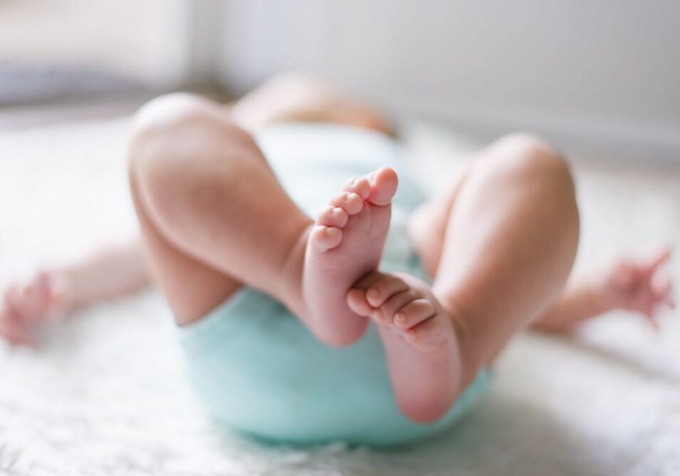 A close-up of a baby lying on a soft surface, wearing a light blue diaper, with legs raised and feet in focus. The background is softly blurred, highlighting the baby's tiny toes and the gentle care that moms trust from all natural products.