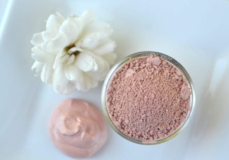 A small glass jar filled with pinkish powder sits on a white surface next to a white flower and a swirl of creamy pink substance—perfect for creating DIY face masks at home.
