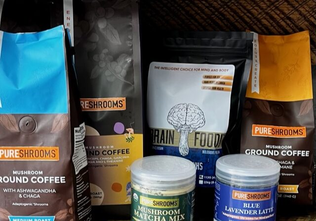 Assorted bags and jars of PureShrooms mushroom coffee and drink mixes are arranged on a wooden shelf, showcasing Mushroom Ground Coffee, Grain Food, Mushroom Matcha Mix, and Blue Lavender Latte with the benefits of mushroom coffee.