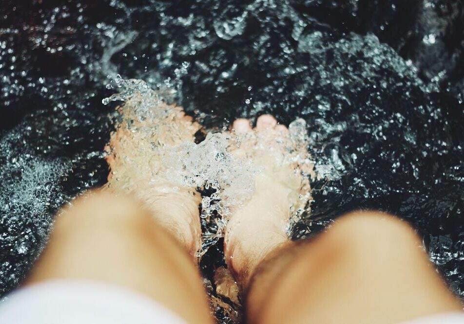 A close-up view of a person’s bare feet submerged in splashing water, perfect for summer foot care, with blurred legs in the foreground and dark water in the background.